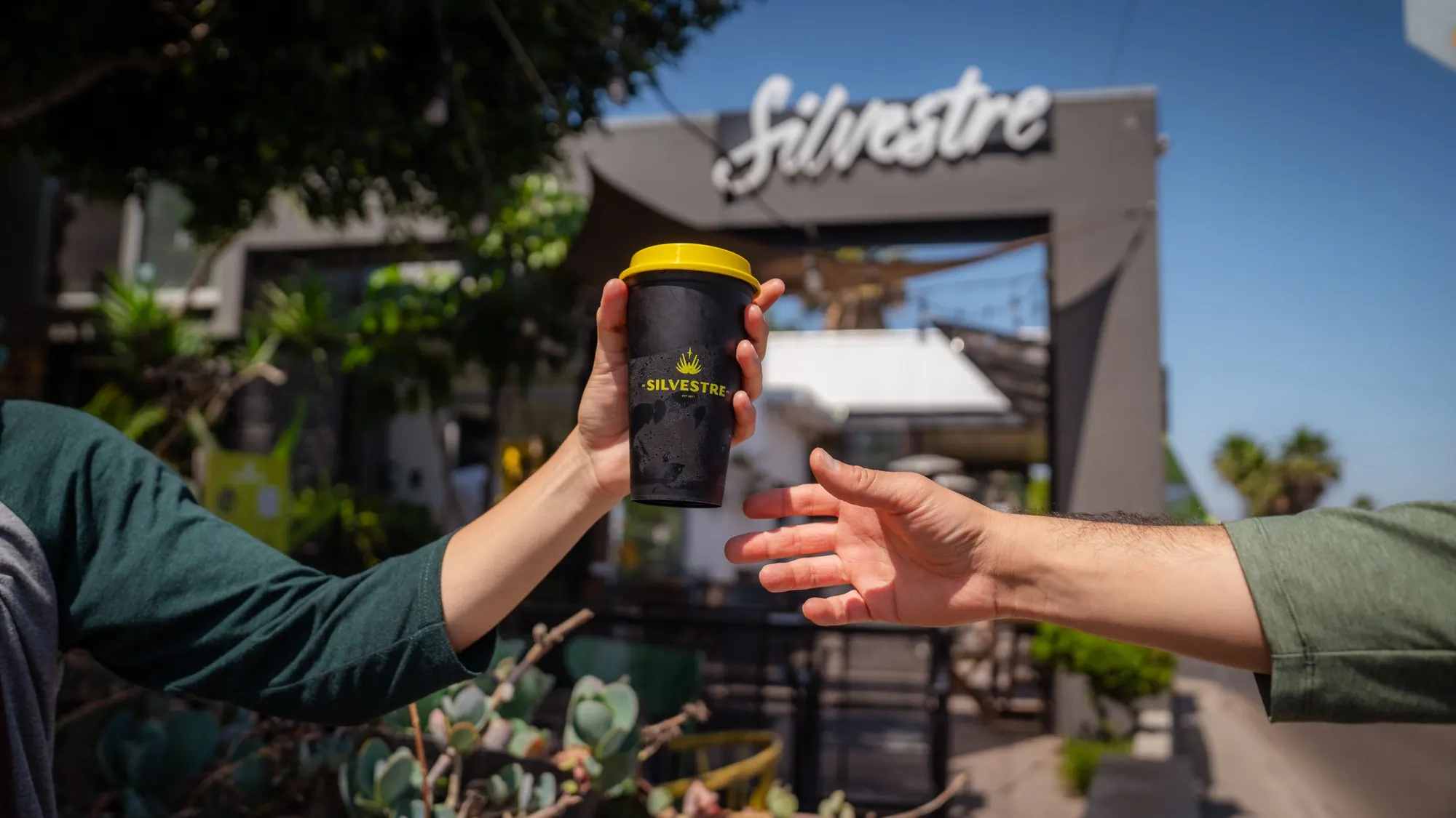 Dos manos se pasan un vaso reusable de Silvestre frente a la fachada del café